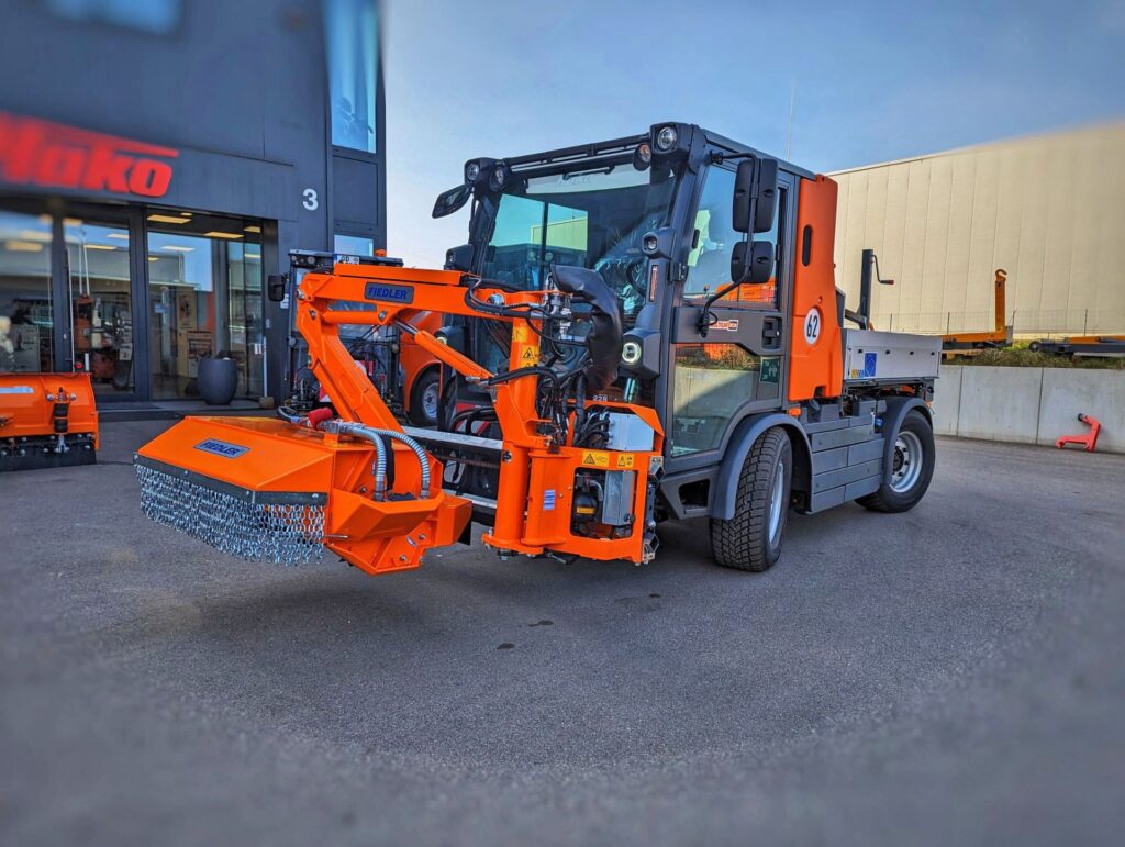 Oranges Kommunalfahrzeug Hako Multicar mit Fiedler Auslegemäher und Kettenvorhang vor einem Firmengebäude.
