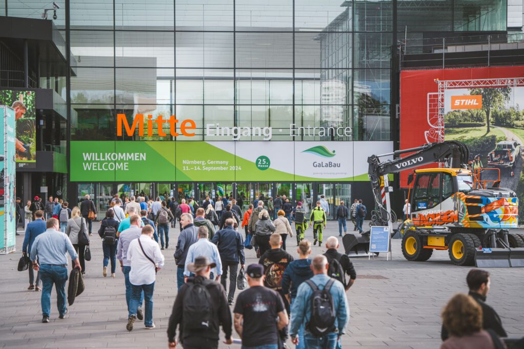 Besucher am Eingang Mitte der GaLaBau Messe Nürnberg mit Volvo-Bagger im Vordergrund und moderner Glasfassade.