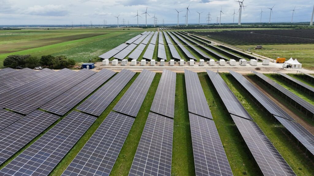 PÜSPÖK realisiert Super-Hybrid-Park in Nickelsdorf 1 Luftaufnahme eines weitläufigen Solarparks mit Photovoltaik-Modulen und Windkraftanlagen im Hintergrund auf grüner Wiese.
