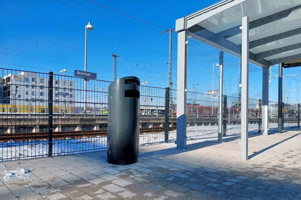 Moderner anthrazitfarbener Mülleimer am Bahnhof Bruchsal neben einem gläsernen Wartebereich bei sonnigem Winterwetter.
