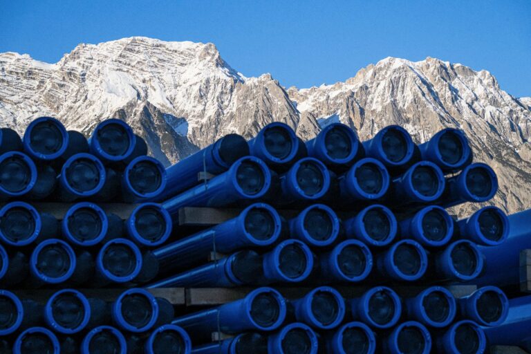 Stapel blauer Rohre auf einer Baustelle vor schneebedeckten Berggipfeln der Alpen unter blauem Himmel.
