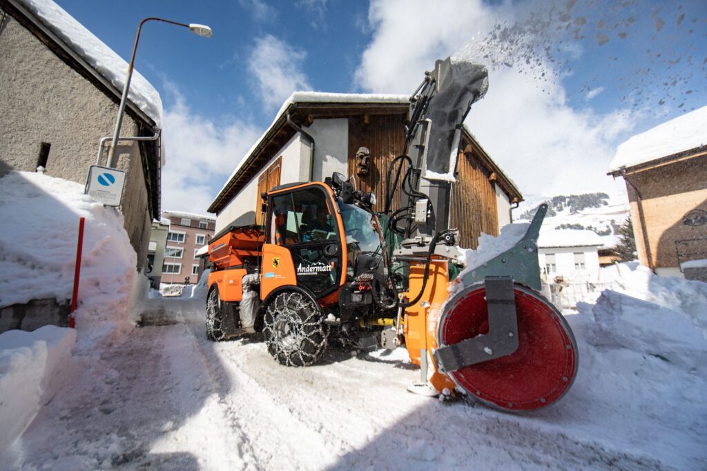 Orangefarbene Schneefräse der Gemeinde Andermatt räumt verschneite Gasse zwischen traditionellen Häusern bei Sonnenschein.