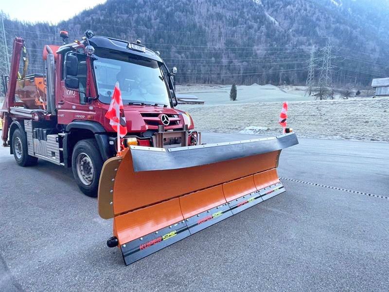 Roter Mercedes-Benz Unimog mit orangefarbenem Schneepflug auf asphaltierter Straße vor bewaldetem Bergpanorama.