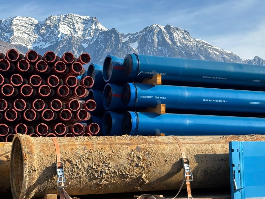 Rohstoff statt Reststoff: Graz setzt auf Kreislaufwirtschaft bei Gussrohren 4 Blaue TRM Gussrohre und braune Muffenrohre gestapelt vor verschneitem Gebirgspanorama der Alpen.