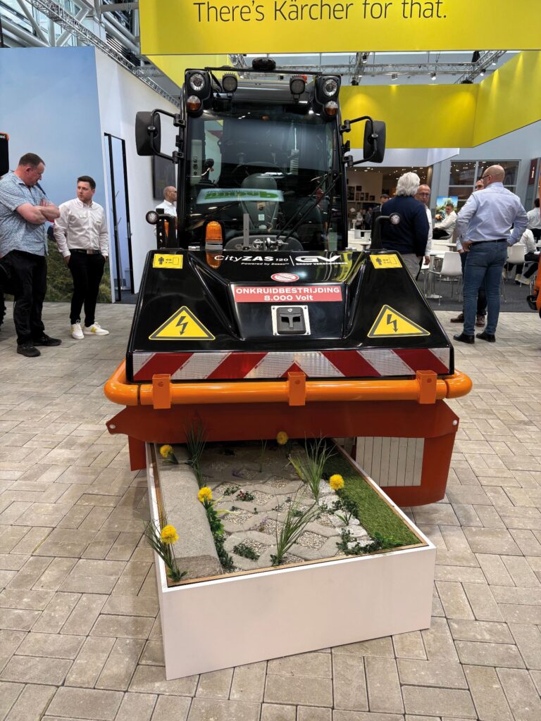 Kärcher präsentiert emissionsfreie Kommunaltechnik auf der Suisse Public 2026 2 CityZAS 120 Kehrmaschine zur elektrischen Unkrautbekämpfung auf einer Messe vor einem Beet mit Löwenzahn.
