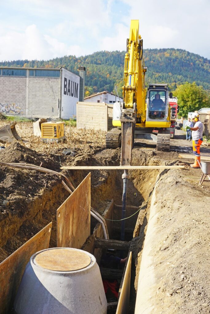 Gelber Kettenbagger mit Hydraulikhammer an einer tiefen Baugrube für Kanalarbeiten mit Betonrohr und Wald im Hintergrund.