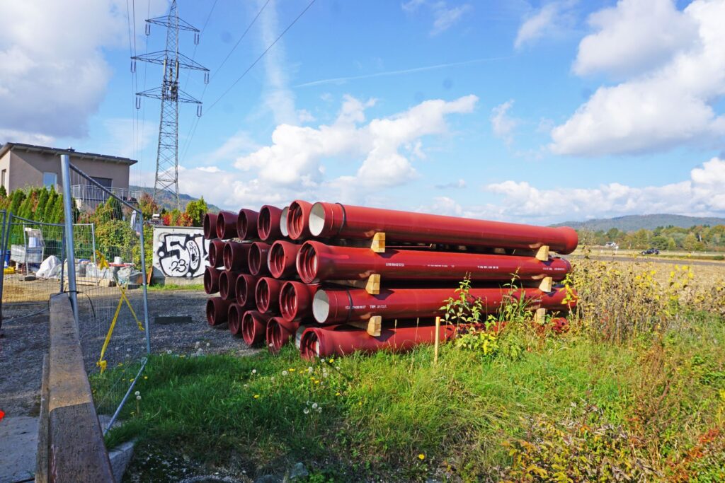 Stapel rote Gussrohre auf Holzlagern im Freien neben einer Baustelle mit Strommast unter blauem Himmel.