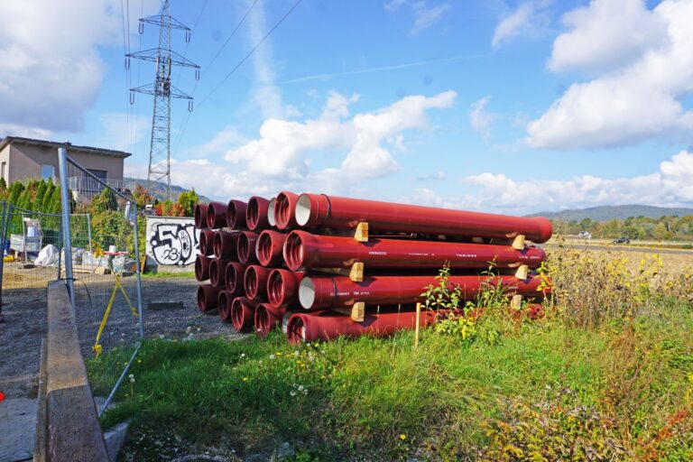 Stapel rote Gussrohre auf Holzlagern im Freien neben einer Baustelle mit Strommast unter blauem Himmel.