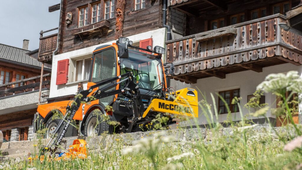 Oranger Holder Geräteträger mit McConnel Power Arm Böschungsmäher vor rustikalem Holzhaus in den Bergen.