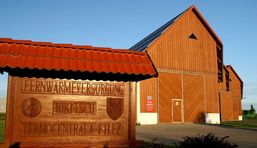 Holzschild der Fernwärme Nikitsch vor einer großen Holzscheune unter blauem Himmel.