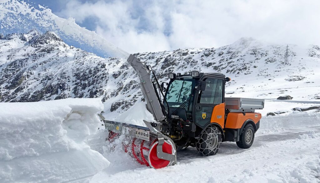 Zaugg entwickelt Bewährtes konsequent weiter: Next Generation Schneepflüge und Concept Blower 13 Orangefarbene Schneefräse von Zaugg räumt eine Hochgebirgsstraße vor schneebedeckter Bergkulisse unter blauem Himmel.