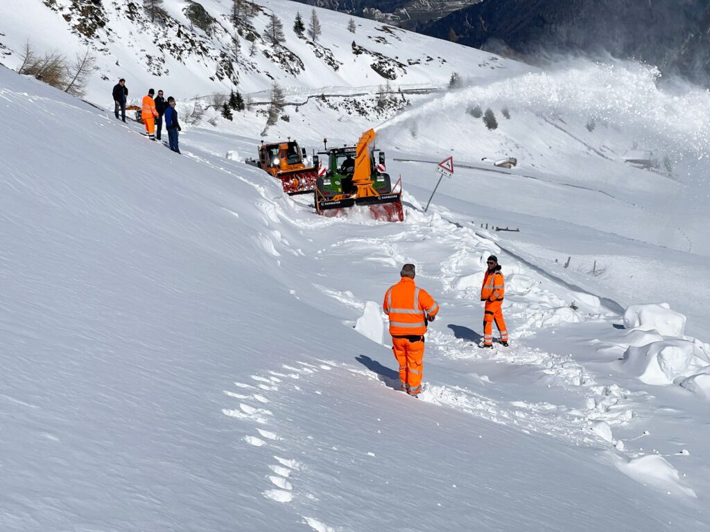 Wenn Schneewände weichen – Kahlbacher räumt Alpenpässe frei 3 Traktoren mit Schneefräsen räumen eine tief verschneite Passstraße im Hochgebirge, beobachtet von Arbeitern in Warnkleidung.