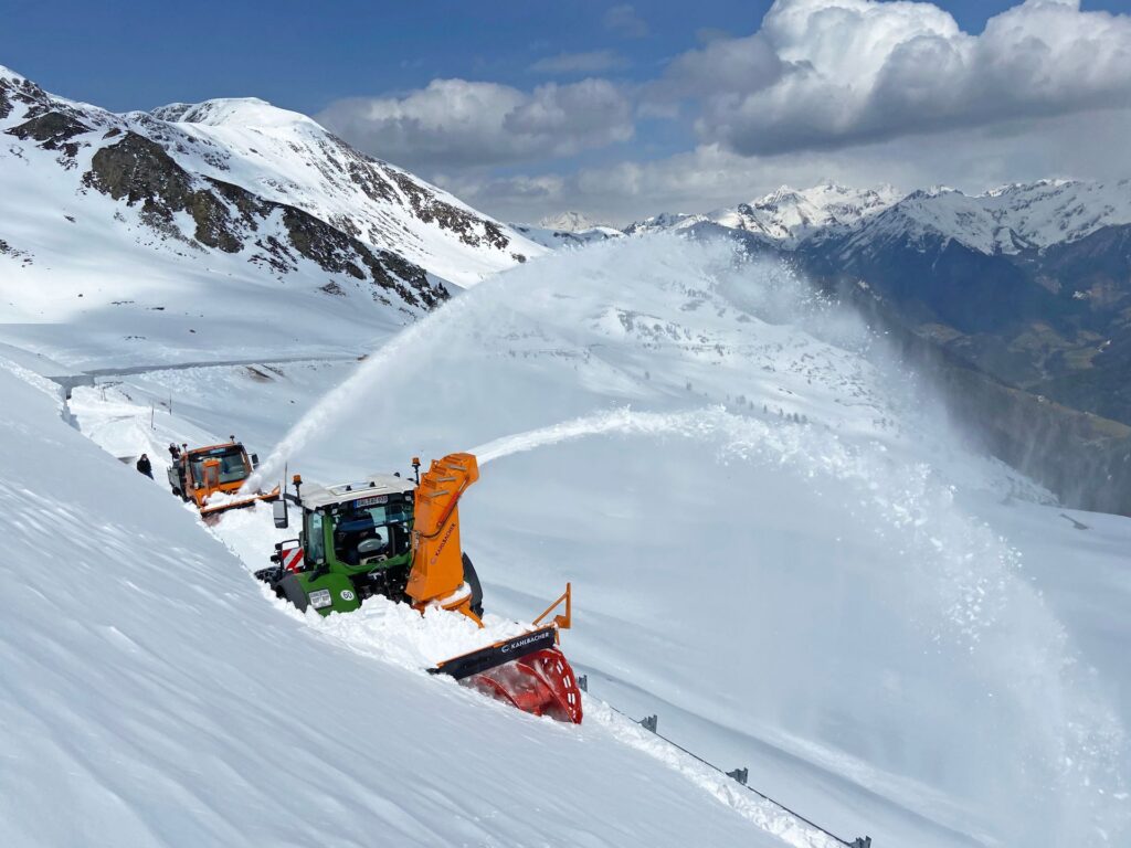 Zwei Schneefräsen räumen eine tief verschneite Passstraße im Hochgebirge vor schneebedeckten Berggipfeln.