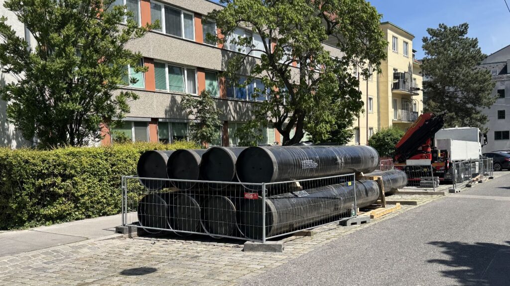 Wien stärkt das Rückgrat seiner Wasserversorgung 5 Gestapelte schwarze Gussrohre hinter Baustellengittern auf einem Gehweg an einer Wohnstraße mit LKW-Kran im Hintergrund.