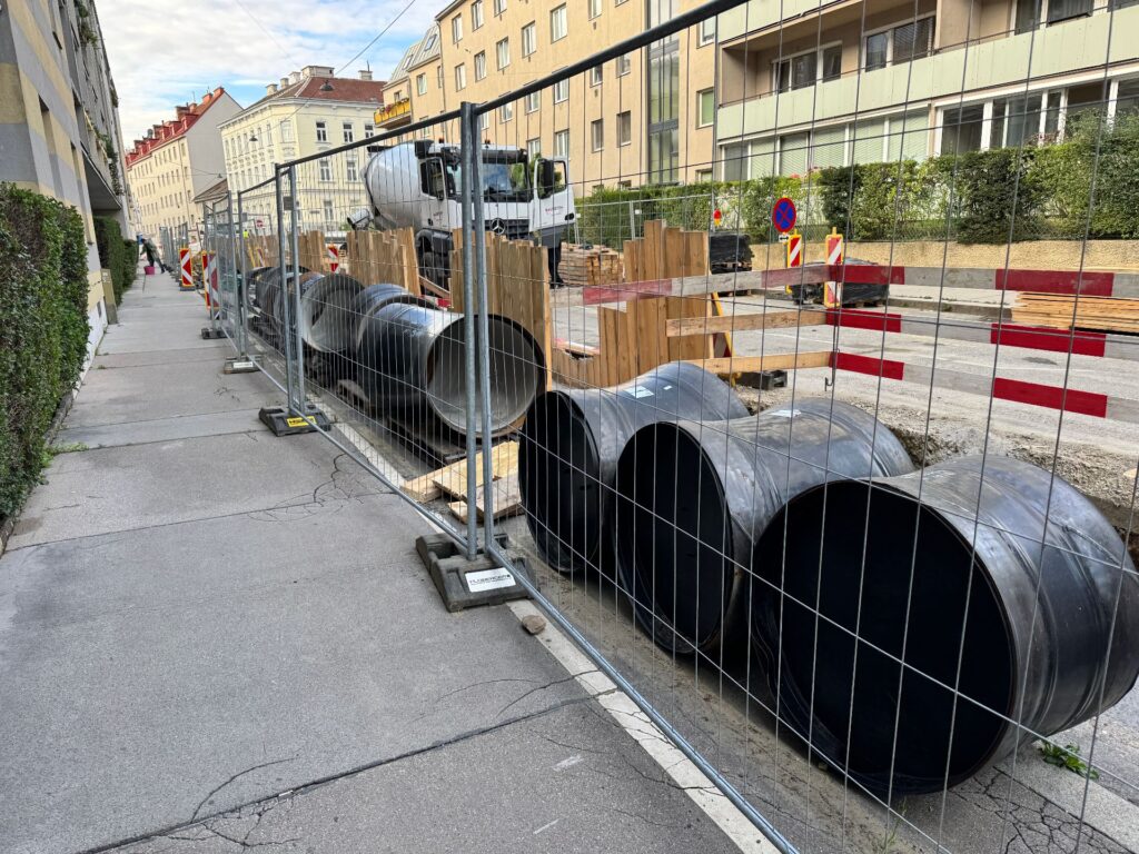 Wien stärkt das Rückgrat seiner Wasserversorgung 4 Große schwarze Rohrsegmente hinter Bauzaun an einer städtischen Baustelle mit Betonmischer im Hintergrund.
