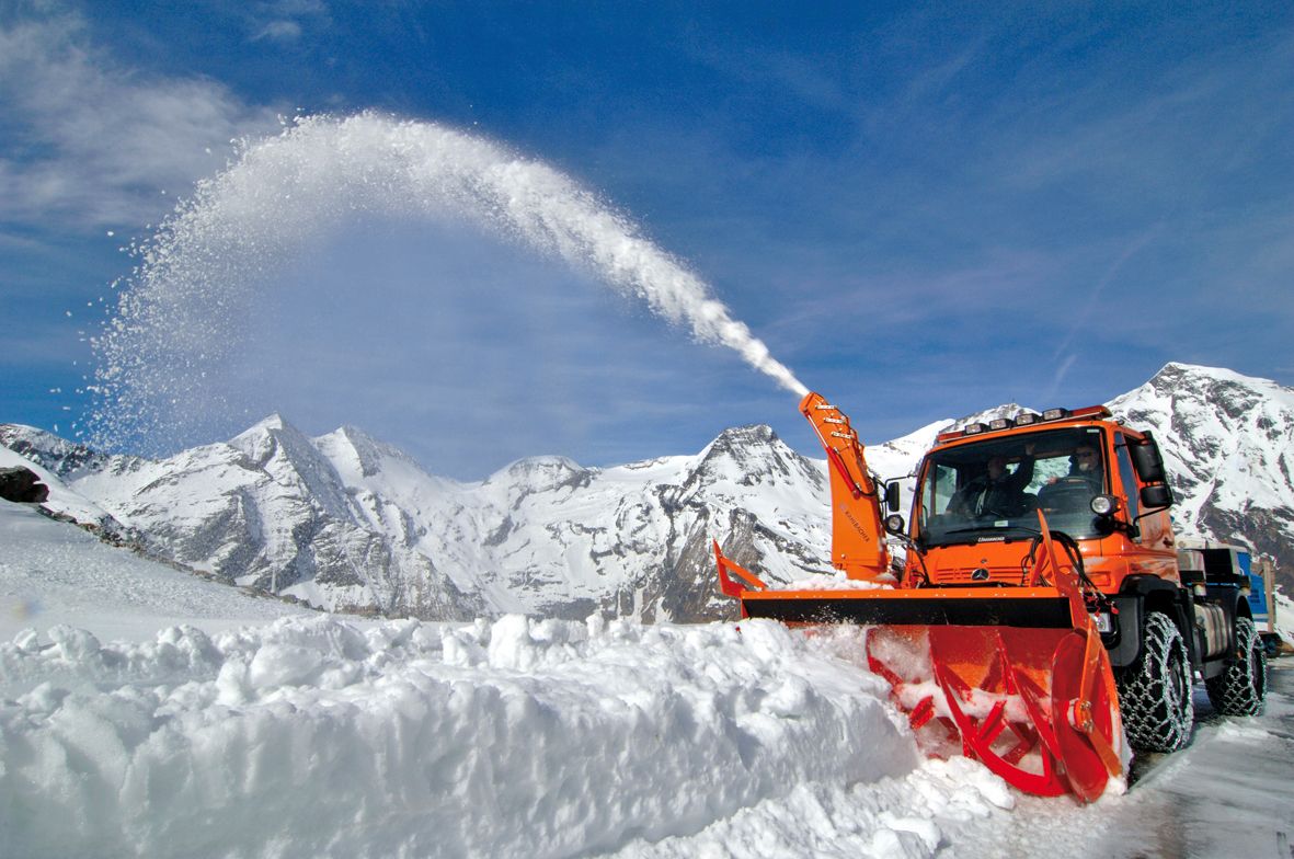 Oranger Unimog mit KFS 950 Schneefrässchleuder wirft hohen Schneebogen vor verschneiter Alpenkulisse und blauem Himmel aus.