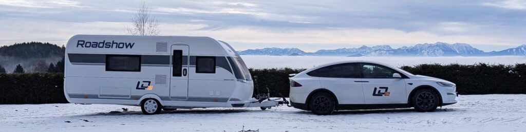 LD2 bringt Vermessung und GIS direkt in die Gemeinde 2 Weißes Tesla Model X zieht LD2 Wohnwagen durch eine verschneite Berglandschaft mit Alpen-Panorama im Hintergrund.