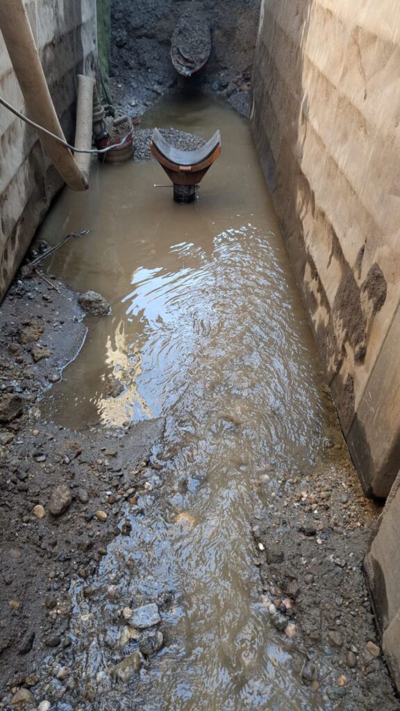 Schlammiges Wasser fließt in einem tiefen Graben mit Verbauwänden an einer Rohrleitung einer Baustelle entlang.