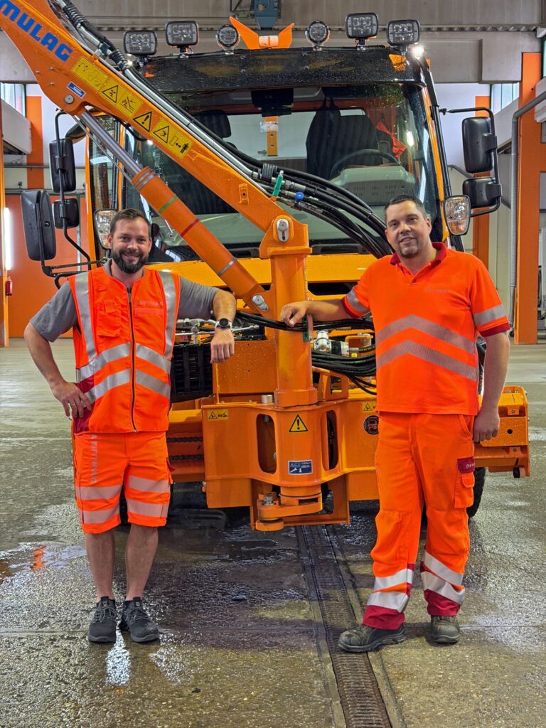 Zwei ASFINAG-Mitarbeiter in Warnkleidung stehen lächelnd vor einem orangefarbenen Unimog mit Mulag-Mähgerät bei Pappas.