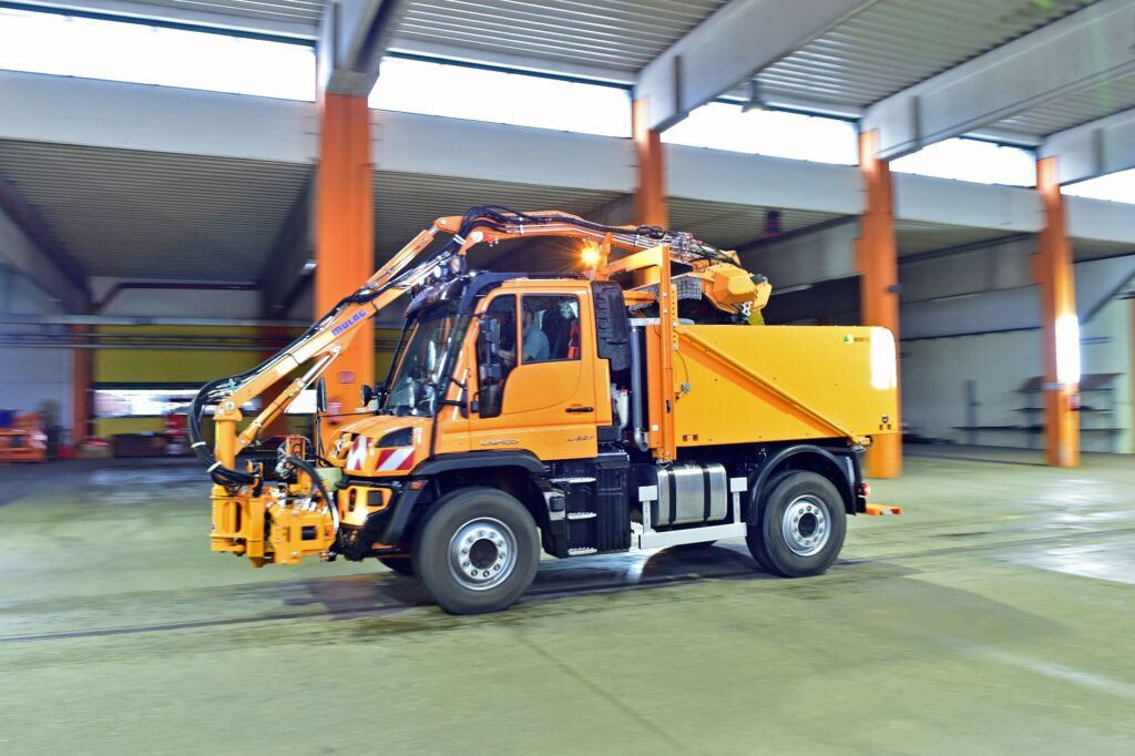 Oranger Mercedes-Benz Pappas Unimog der ASFINAG mit Ecotech-Aufbau und Mulag-Mähausleger in einer Wartungshalle.