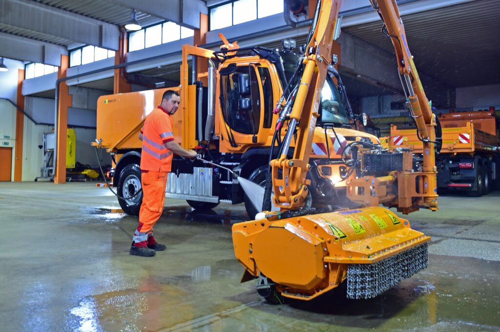Mitarbeiter reinigt orangen Pappas Unimog mit Ecotech-Aufbau und ASFINAG-Mähgerät per Hochdruckreiniger in einer Halle.