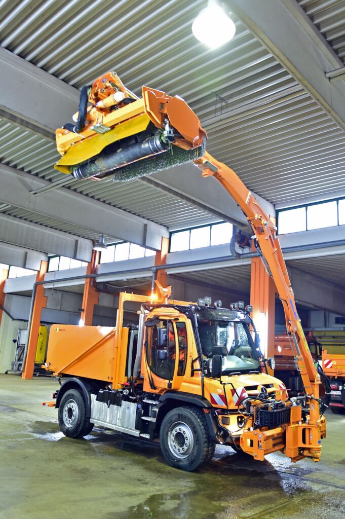 Oranger Pappas Unimog der Asfinag mit Ecotech-Aufbau und erhobenem Mäharm in einer Wartungshalle.