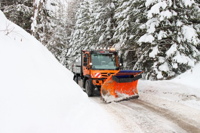 Oranger RW Bau Unimog mit Schneepflug bei der Schneeräumung auf einer Waldstraße im tiefen Winter.