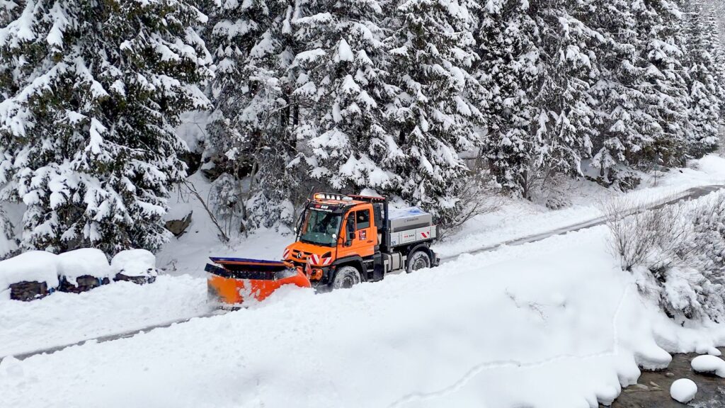 Stark im Steilhang: Sicher unterwegs im Paznauner Winterdienst 2 Orangefarbener RW13 Unimog Winterdienst beim Schneeräumen auf einer verschneiten Straße vor tief verschneitem Nadelwald.