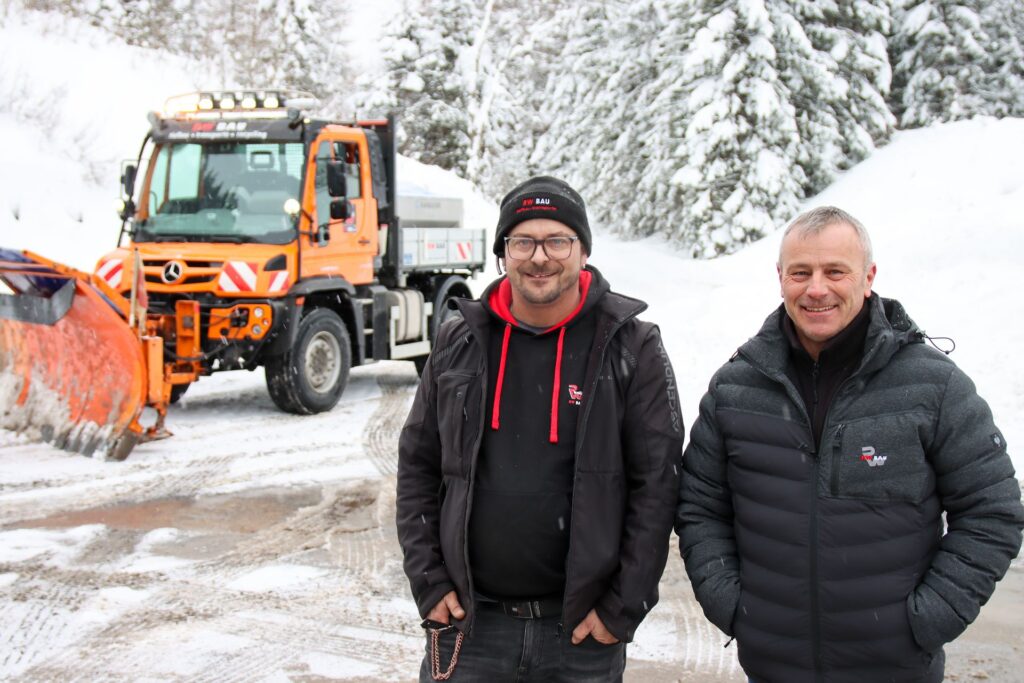 Stark im Steilhang: Sicher unterwegs im Paznauner Winterdienst 1 Zwei lächelnde Mitarbeiter der RW Bau vor einem Mercedes Unimog Winterdienst-Fahrzeug mit Schneepflug im verschneiten Wald.