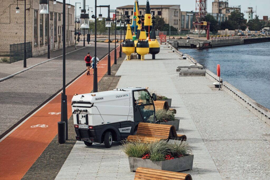 Bucher CityCat VR17e: Vollelektrische Kompaktkehrmaschine für den urbanen Einsatz 1 Kompakte Bucher CityCat VR17e Kehrmaschine demonstriert engen Wendekreis auf einer Uferpromenade neben einem Radweg.