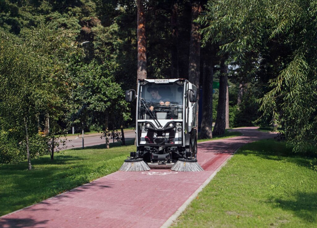 Bucher CityCat VR17e: Vollelektrische Kompaktkehrmaschine für den urbanen Einsatz 2 Kompakte elektrische Kehrmaschine CityCat VR17e reinigt einen roten Radweg im Park bei Sonnenschein.