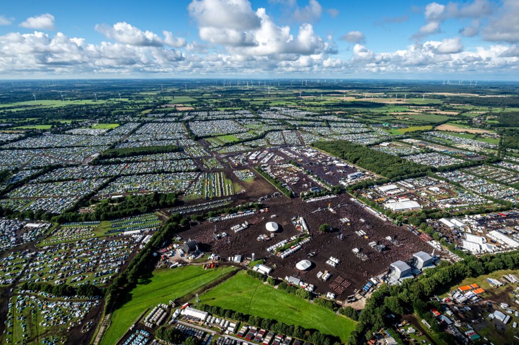 Luftaufnahme des Wacken Open Air Festivals mit riesigem Campingplatz, Hauptbühnen und Besuchermengen vor flacher Landschaft.