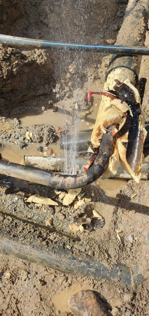 Wasser spritzt aus einer beschädigten, isolierten Rohrleitung in einer erdigen Baugrube; akuter Rohrbruch und Wasserschaden.