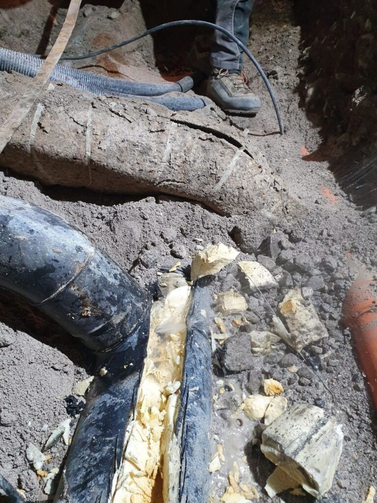Freigelegte, beschädigte Fernwärmeleitung mit gelber Isolierung in einer Baustelle im Erdreich neben weiteren Rohren.