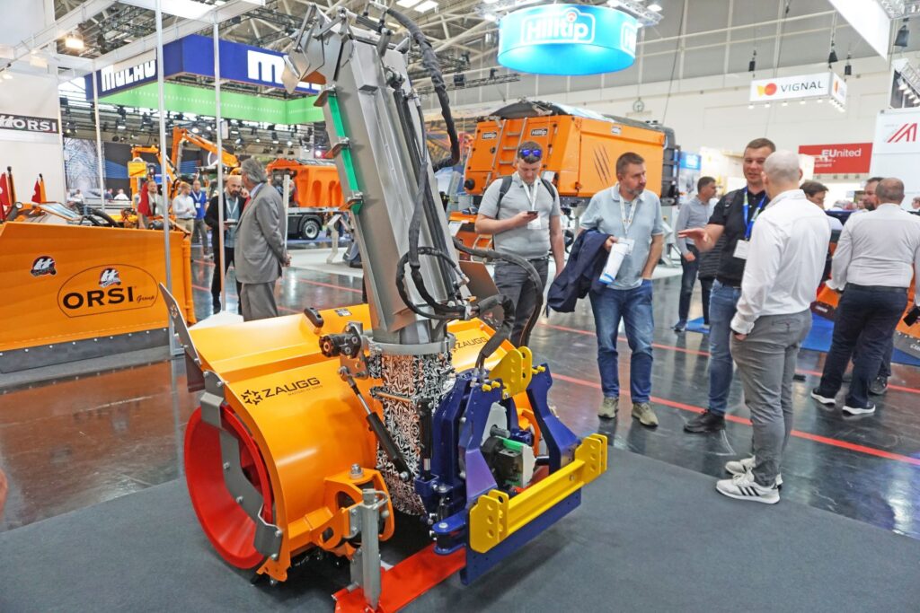 Orange Zaugg Schneefräse auf einer Messe, umgeben von Besuchern und weiteren Winterdienstfahrzeugen in einer Messehalle.