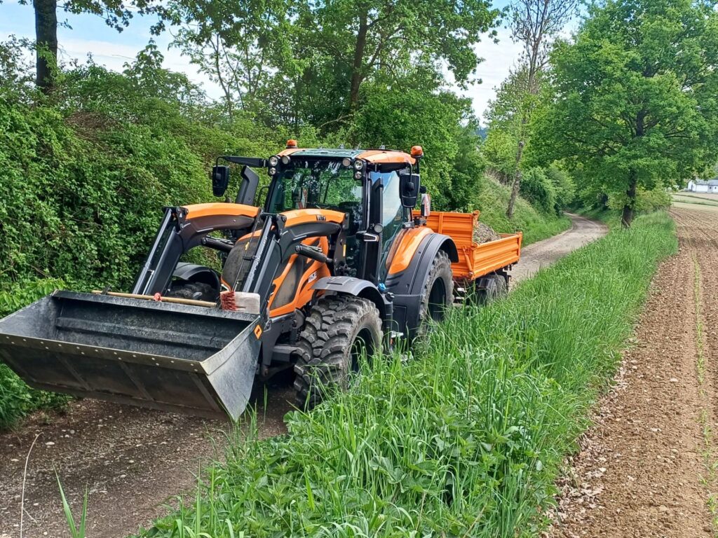 Mauch punktet mit Kompetenz und Verlässlichkeit im Kommunalbereich 2 Oranger Valtra Traktor mit Frontlader-Schaufel und Anhänger auf einem Feldweg neben grünen Wiesen und Bäumen.