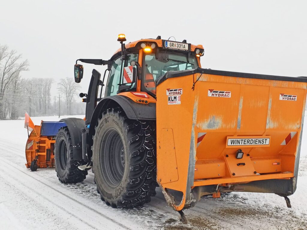 Mauch punktet mit Kompetenz und Verlässlichkeit im Kommunalbereich 1 Oranger Winterdienst-Traktor mit Hydrac Schneepflug und Salzstreuer im Einsatz auf verschneiter Straße.