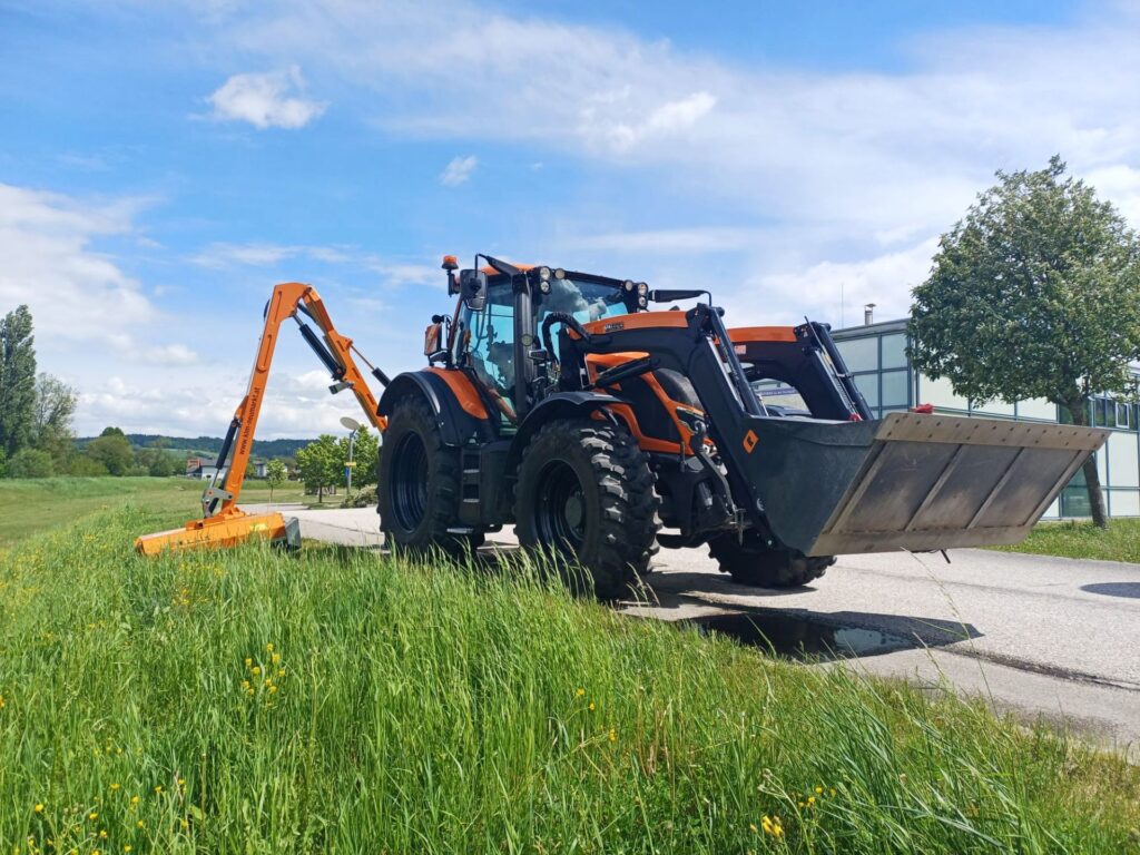 Oranger Valtra Traktor mit Frontlader und Auslegemäher bei der Landschaftspflege auf einer grünen Wiese unter blauem Himmel.