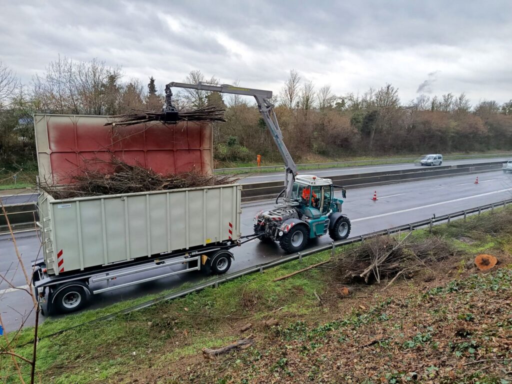 Pfanzelt Systemschlepper lädt mit einem Forstkran Gehölzschnitt an einer Autobahn in einen Containeranhänger.