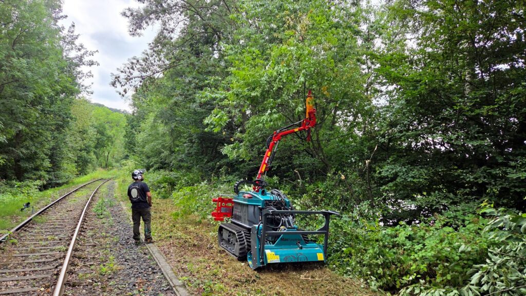 Pfanzelt Forstfräse bei der Gehölzpflege an Bahngleisen; ferngesteuerte Raupe stutzt Äste mit ausfahrbarem Greifarm.