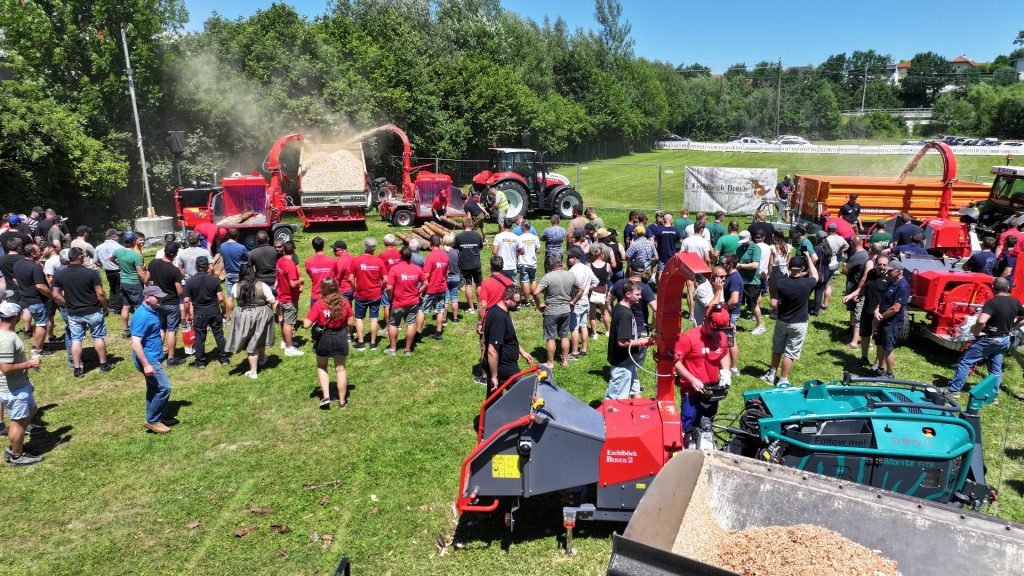 Publikum beobachtet Vorführung roter Holzhacker und Traktoren auf einer Wiese bei einer Forsttechnik-Messe im Freien.