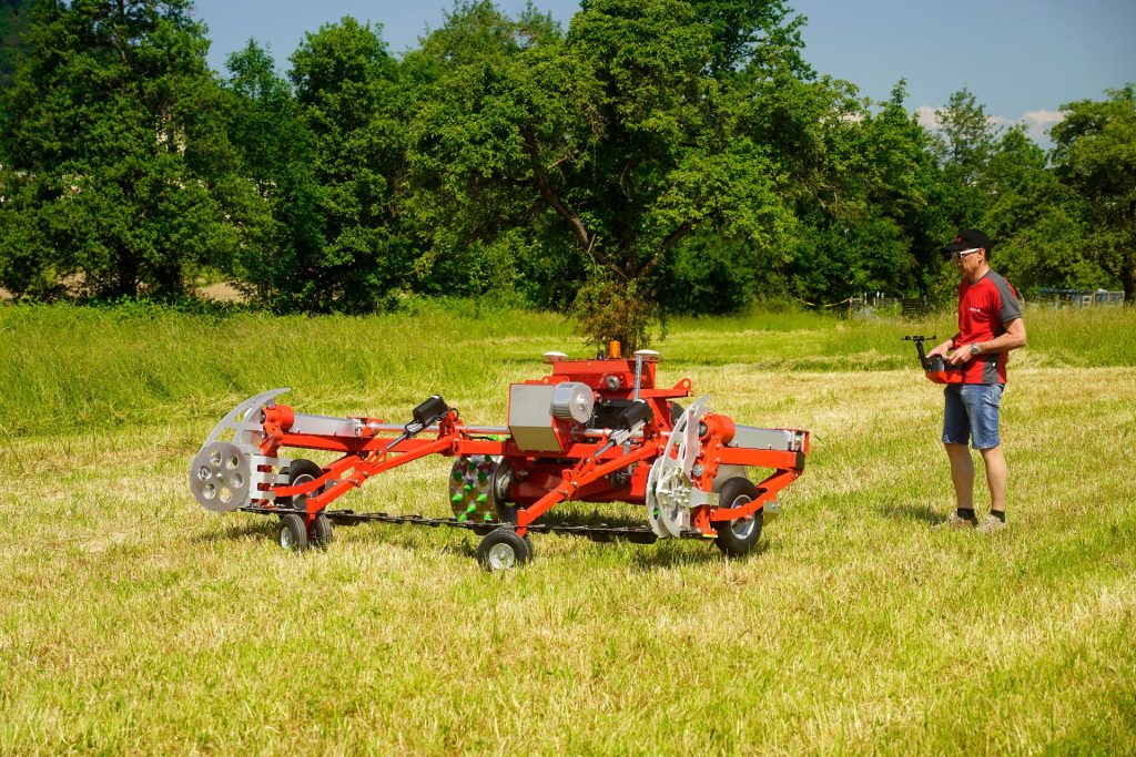 100 Jahre Rapid: Vom Motormäher zur vernetzten Kommunaltechnik 5 Mann steuert ferngesteuerte rote Landmaschine mit Mähwerk auf einer grünen Wiese unter blauem Himmel.