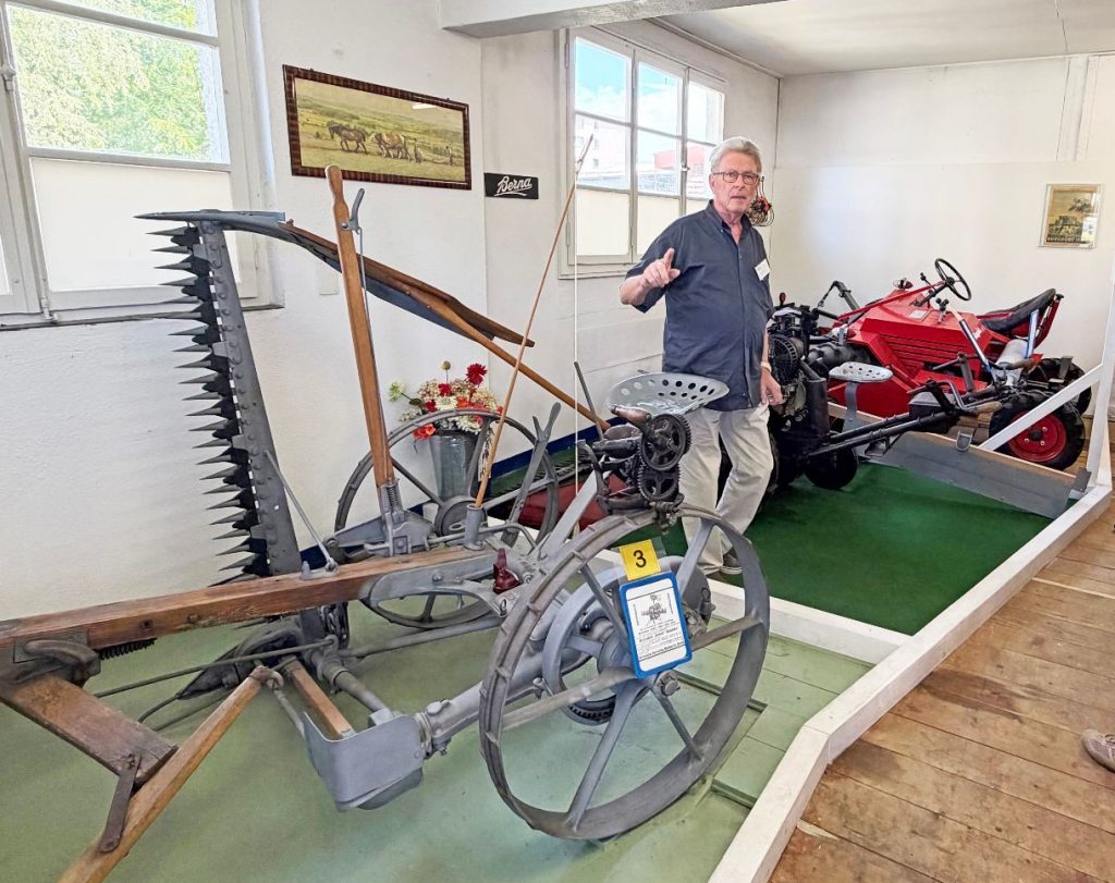 100 Jahre Rapid: Vom Motormäher zur vernetzten Kommunaltechnik 1 Mann erklärt Besuchern im Museum historische Landmaschinen wie einen Rapid Motormäher und einen grauen Balkenmäher.