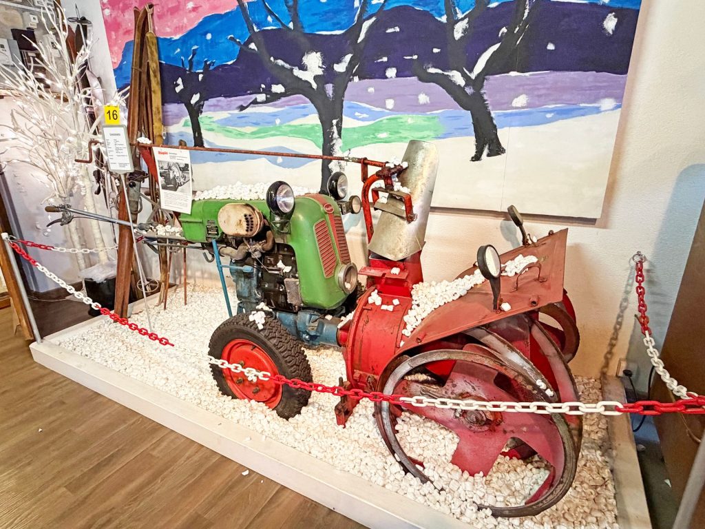 100 Jahre Rapid: Vom Motormäher zur vernetzten Kommunaltechnik 2 Historische Rapid Schneefräse in Grün und Rot als Museumsexponat vor einem Wintergemälde auf künstlichem Schnee.
