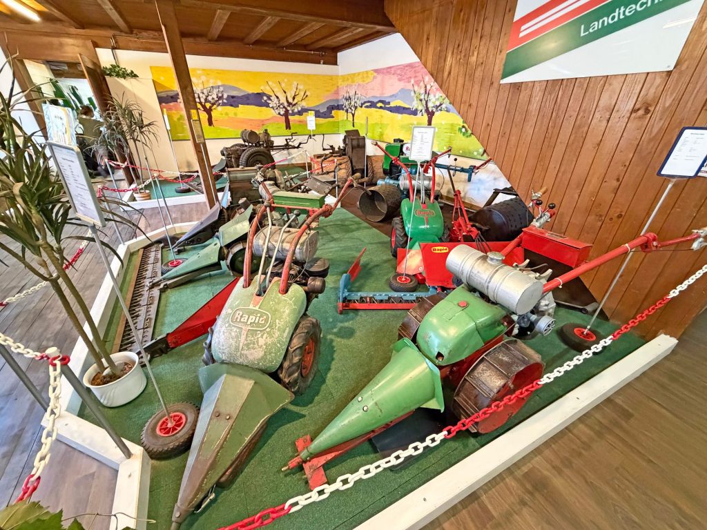 100 Jahre Rapid: Vom Motormäher zur vernetzten Kommunaltechnik 3 Historische Rapid Motormäher und Landmaschinen in einer Ausstellung mit Holzverkleidung und Wandgemälde.