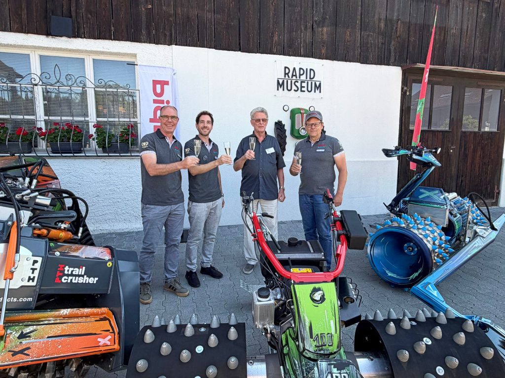 Vier Männer stoßen mit Sektgläsern vor dem Rapid Museum an, umgeben von Einachsschleppern und einem PTH Trail Crusher.